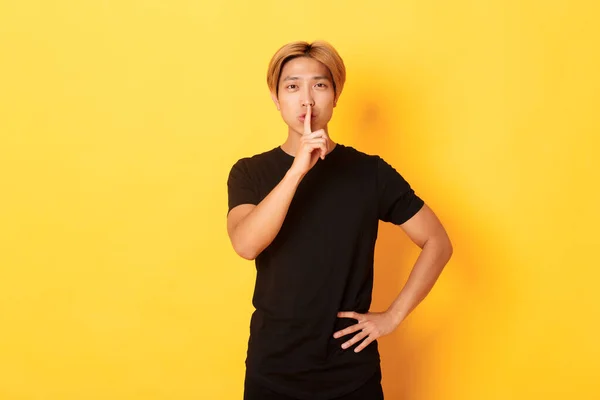 Портрет привабливого блондинки азіатського чоловіка, що стискає, натискає пальцем на губи і лущить, жовтий фон — стокове фото
