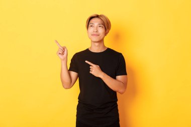 Düşünceli ve memnun Asyalı yakışıklı adam gülümsüyor ve sol üst köşeyi işaret ediyor, sarı arka planın üzerinde duruyor.