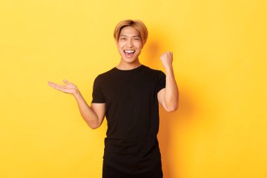 Memnun ve mutlu çekici Koreli adam gülümsüyor, sevinçle yumruk sallıyor, elinde bir şey tutuyor, sarı arka planda duruyor.