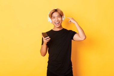 Mutlu Asyalı adam müziği ya da podcast 'i seviyor, kulaklıkları işaret ediyor, akıllı telefon tutuyor, sarı arka planda duruyor.