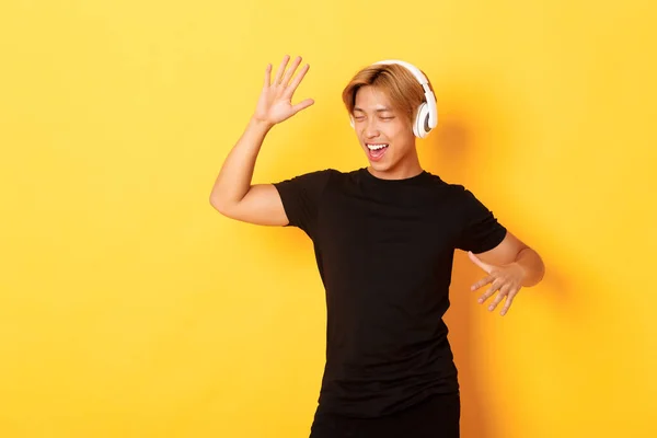 Heureux attrayant asiatique guy avec blond cheveux, chanter le long et danser comme écouter de la musique dans sans fil casque, debout fond jaune — Photo