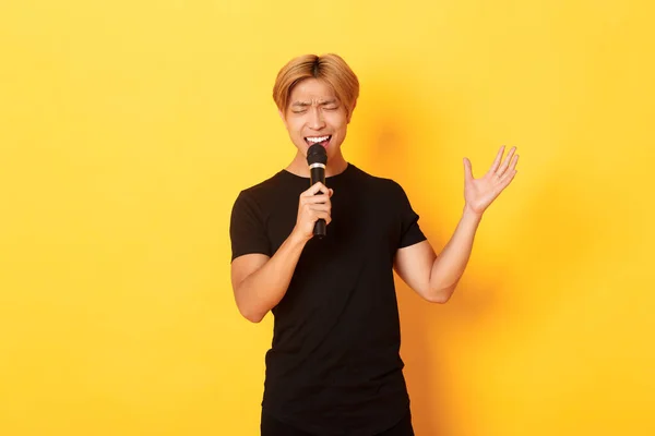 ハンサムなアジア系男性歌手,韓国人男性歌カラオケでマイクで情熱を持って,黄色の背景の上に立って — ストック写真