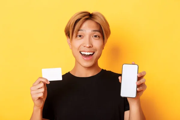 Sorprendido chico asiático feliz mostrando la tarjeta de crédito y la pantalla del teléfono inteligente, sonriendo fascinado, de pie fondo amarillo — Foto de Stock