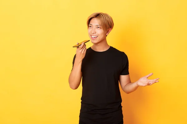 Портрет стильного корейського хлопця зі світлим волоссям запис голосового повідомлення на мобільному телефоні, тримає смартфон біля рота і говорить, стоячи жовтим фоном — стокове фото
