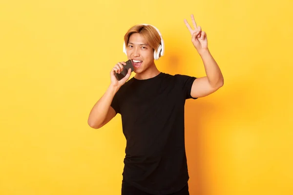 Саші гарний азіатський хлопець в навушниках, грає в караоке-додаток, співає в мікрофон мобільного телефону, демонструє жест миру, стоїть жовтий фон — стокове фото