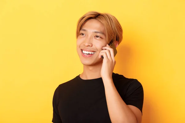 Närbild av snygg bekymmerslös asiatisk kille med ljust hår, talar på mobiltelefon och ler glad, stående gul bakgrund — Stockfoto