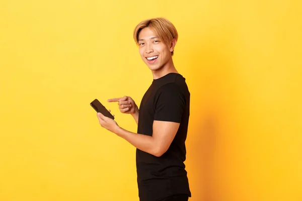 Portrét potěšené usmívající se pohledný asijský muž, stojící v profilu a ukazující prstem na smartphone, doporučuji app, stojící žluté pozadí — Stock fotografie
