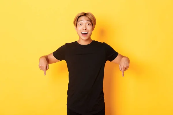 Портрет збудженого усміхненого азіатського хлопця з світлим волоссям, вказуючи пальці вниз, показуючи прапор і виглядаючи щасливим, жовтим фоном — стокове фото
