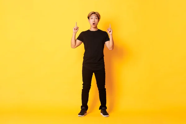 Full-length van onder de indruk aziatische man in zwarte kleren, hijgend verbaasd en wijzende vingers omhoog, gele achtergrond — Stockfoto