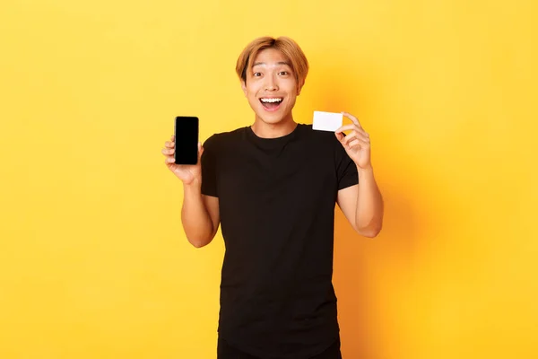 Retrato de animado feliz asiático homem mostrando tela do telefone móvel e cartão de crédito com sorriso alegre, de pé fundo amarelo — Fotografia de Stock