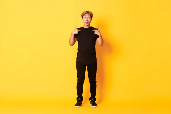 Full-length překvapený atraktivní korejský blonďák v černém oblečení ukazuje na sebe dojem, žluté pozadí — Stock fotografie