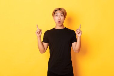 Siyah tişörtlü afallamış Asyalı adamın portresi, ağzı açık büyülenmiş, parmakları havada, sarı arka planda.