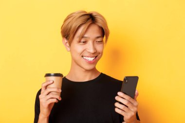 Cep telefonu kullanan ve kahve içen yakışıklı Asyalı erkek öğrencinin yakın çekimi. Adam akıllı telefondan mesajlaşıyor, sarı arka planda duruyor.