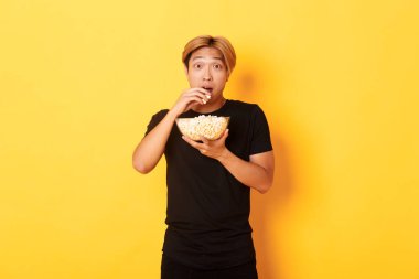 Heyecanlı ve şaşırmış Asyalı adam ilginç bir film izliyor ve kaseden patlamış mısır yiyor, sarı arka planda duruyor.