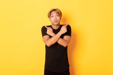 Kararsız yakışıklı adam şaşırmış ve parmaklarını yana doğru gösteriyor, sarı arka planda duruyor.