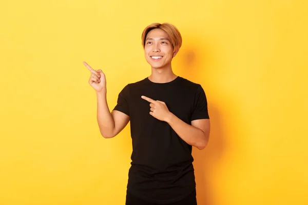 Portrét pohledného korejského blonďáka, ukazující prsty v levém horním rohu a spokojeně se usmívající, žluté pozadí — Stock fotografie