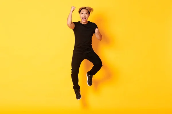 Pleine longueur de heureux attrayant asiatique guy en noir vêtements sautant et célébrant la victoire, atteindre objectif, debout fond jaune — Photo