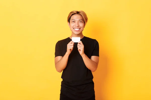 Emocionado chico asiático feliz sonriendo y mostrando tarjeta de crédito, de pie fondo amarillo — Foto de Stock