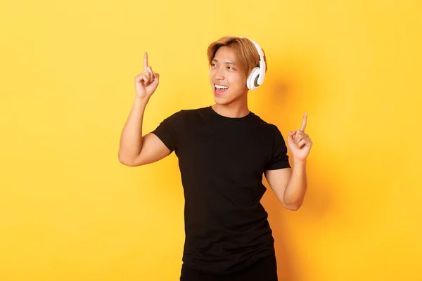 Joyful handsome asian guy with blond hair, singing along and dancing as listening to music in wireless headphones, standing yellow background