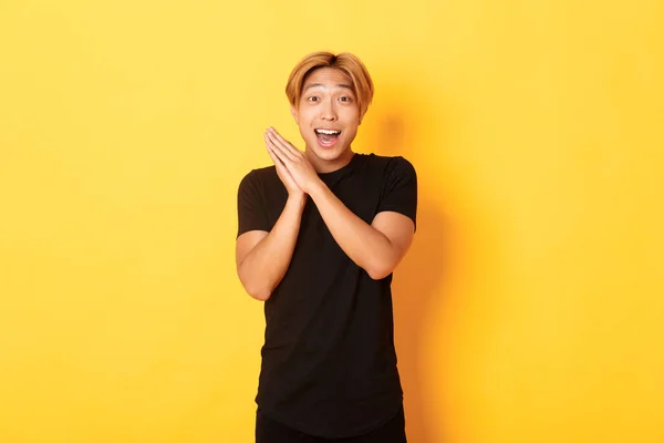 Retrato de feliz e surpreso asiático homem palmas mãos e sorrindo encantado, de pé fundo amarelo — Fotografia de Stock