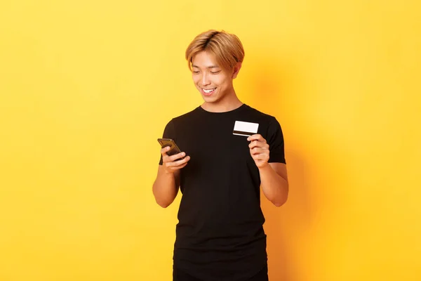 Sonriente guapo asiático hombre pagar por la compra en línea, compras en Internet, la celebración de teléfono inteligente y tarjeta de crédito, fondo amarillo — Foto de Stock