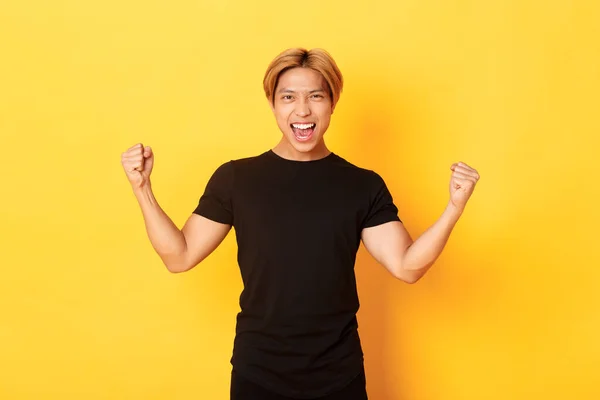 Retrato de feliz atraente asiático homem regozijando, levantando as mãos para cima e gritando sim, de pé satisfeito sobre fundo amarelo — Fotografia de Stock