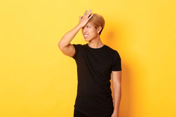 Rozrušený a problémový asijský chlap chňapnout čelo zapomnětlivý, stojící nad žlutým pozadím — Stock fotografie