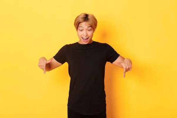 Portrait of excited and happy asian guy with blond haircut, pointing and looking down amused, standing yellow background — Stock Photo, Image