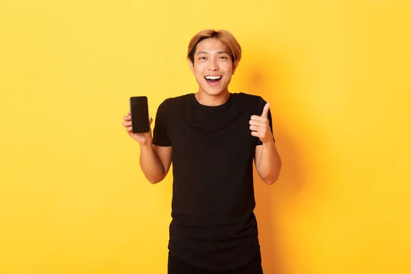 Портрет задоволеного усміхненого азіатського хлопця, який показує екран смартфона і великі пальці задоволені додатком, стоячи жовтим тлом — стокове фото