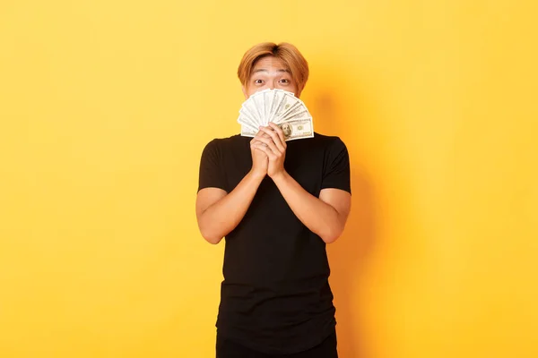 Взволнованный счастливый блондин азиатский парень радуется выигрышу, держит деньги и выглядит счастливым, стоя на желтом фоне — стоковое фото