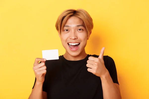 행복하고 만족 해 하는 이 시안잘생긴 남자의 클로즈업 신용 카드와 엄지 사진을 보여 주며 웃으며 노랗게 서 있는 모습 — 스톡 사진