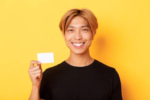 Närbild av snygg nöjd asiatisk kille med ljust hår, leende glad och visar kreditkort över gul bakgrund — Stockfoto