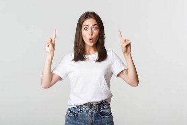 Şaşırmış ve etkilenmiş esmer kız şaşkın şaşkın bakıyor ve parmakla işaret ediyor.