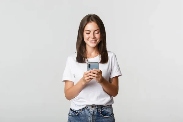 Улыбаясь привлекательная девушка с помощью мобильного телефона и глядя на экран приятно — стоковое фото