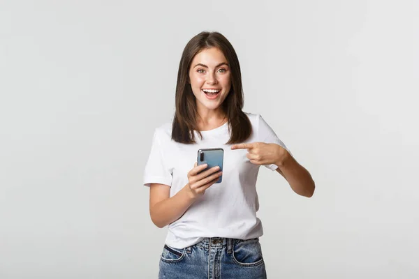 Eccitato e contento ragazza bruna puntando il dito contro lo schermo dello smartphone — Foto Stock