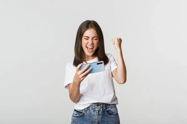 Glädjande söt flicka spelar mobiltelefon spel och knytnäve pump, nöjd med vinna — Stockfoto