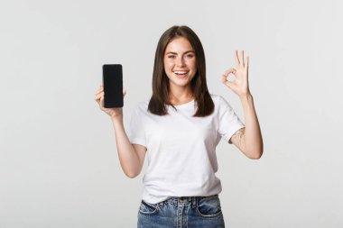 Sevinçli esmer kız akıllı telefon ekranı gösteriyor ve onaylanmış jest yapıyor.