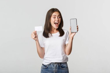 Heyecanlı ve şaşırmış güzel kız ekranda kredi kartı ve cep telefonu bankacılık uygulaması gösteriyor.