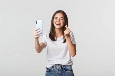 Mutlu, güzel, genç bir kadın kalp hareketi yapıyor ve akıllı telefondan selfie çekiyor, kaygısızca gülüyor.