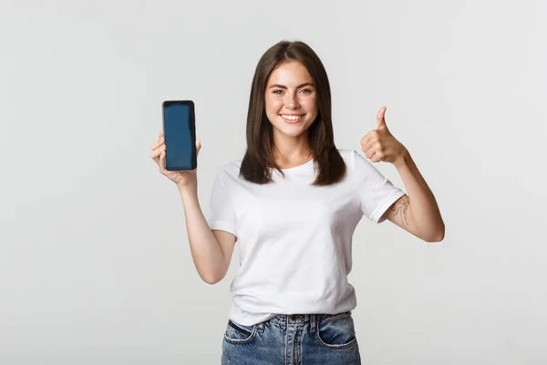 Nöjd leende attraktiv kvinna visar smartphone skärm och tummen upp — Stockfoto