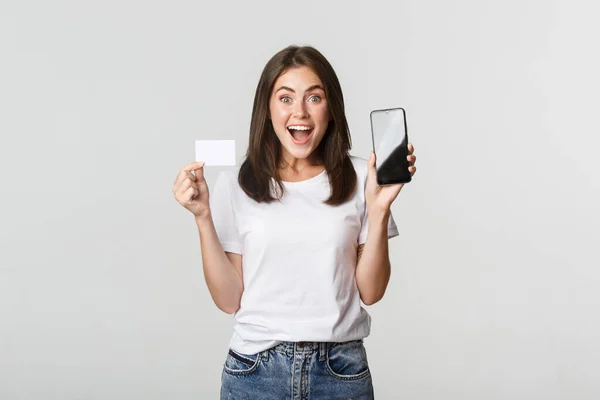 興奮し、驚いてかわいい女の子が画面上でクレジットカードや携帯電話の銀行アプリを表示 — ストック写真