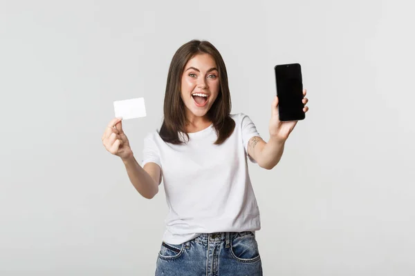 Attraktiv leende flicka ser nöjd och visar kreditkort, mobiltelefon skärm — Stockfoto