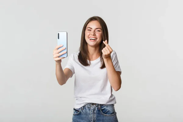 Feliz hermosa mujer joven mostrando el gesto del corazón y tomar selfie en el teléfono inteligente, riendo sin preocupaciones — Foto de Stock