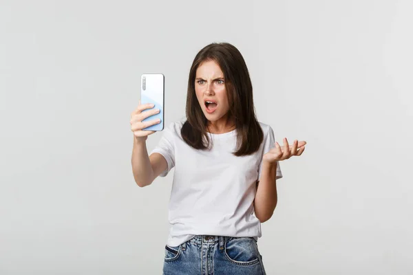 Mujer joven frustrada discutiendo en videollamada, sosteniendo el teléfono inteligente, de pie fondo blanco —  Fotos de Stock