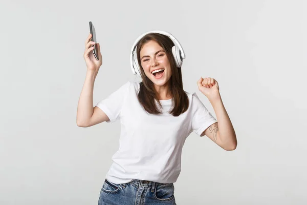 stock image Happy beautiful brunette girl dancing and listening music in wireless headphones, holding smartphone