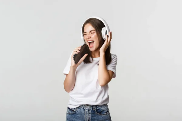 Mulher feliz atraente jogando aplicativo de karaoke, cantando no smartphone, usando fones de ouvido sem fio — Fotografia de Stock