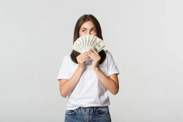 Emocionado chica de ensueño sosteniendo el dinero y mirando esquina superior izquierda, pensando sobre fondo blanco — Foto de Stock