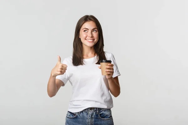 Menina morena sorridente atraente olhando satisfeito, beber café e mostrar polegares para cima, recomendar café — Fotografia de Stock