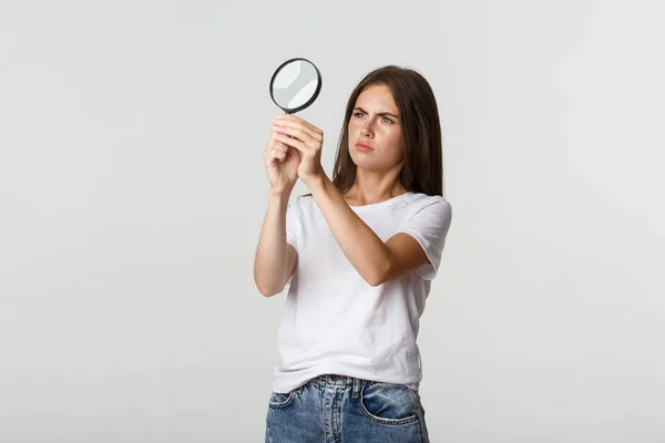 Serieus uitziende aantrekkelijke jonge vrouw op zoek naar iets, op zoek door vergrootglas, witte achtergrond — Stockfoto