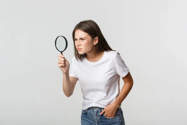 Serieus uitziende aantrekkelijke jonge vrouw op zoek naar iets, op zoek door vergrootglas, witte achtergrond — Stockfoto